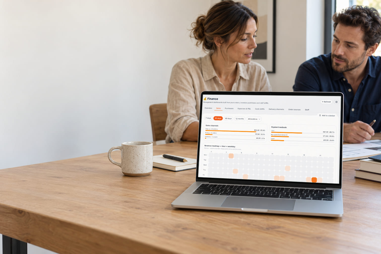 A business owner and a colleague review their results; the laptop shows the real financial overview